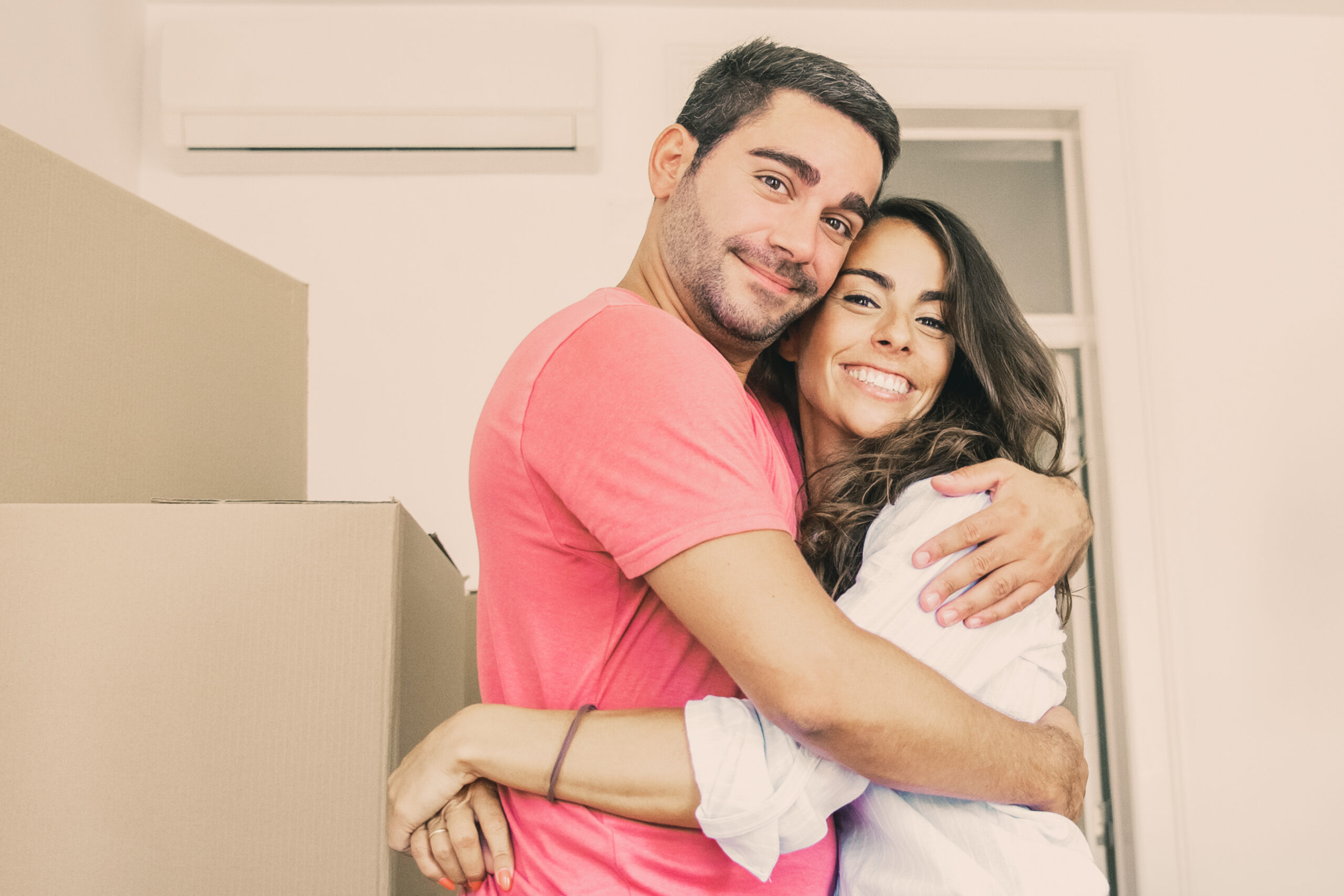Cheerful couple moving into new flat, standing among carton boxes and hugging. Medium shot. New home concept